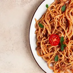Talerz spaghetti bolognese z pomidorkiem i bazylią na jasnym tle.