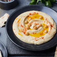 Kremowy hummus na talerzu, polany oliwą, posypany papryką i ziarnami ciecierzycy. Obok kawałki pity i czosnek.