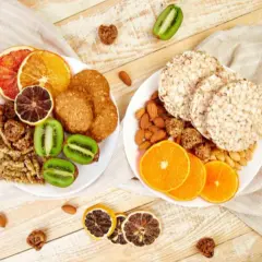 Dwa talerze z przekąskami: ciastka, suszone owoce, kiwi, migdały i wafle ryżowe.