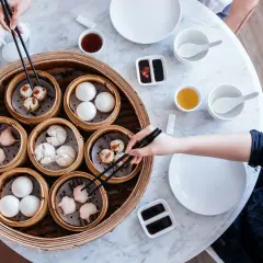 Ręce sięgają po pyszne dim sum z bambusowego parownika. Na stole stoją miseczki z sosami i herbatą.