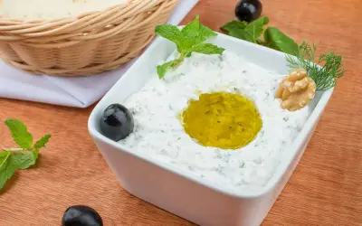 Jak zrobić tzatziki? Kremowy dip jogurtowy z oliwą, miętą, orzechami i oliwkami. Obok koszyk z pitą.