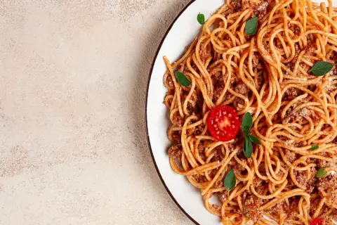 Talerz spaghetti bolognese z pomidorkiem i bazylią na jasnym tle.