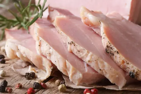 Super delikatny i soczysty schab gotowany, pokrojony w plastry, z gałązką rozmarynu i ziarnami pieprzu na drewnianym tle.