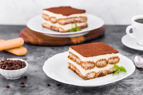 Pyszne tiramisu z warstwami kremu i biszkoptów do tiramisu, posypane kakao. Obok filiżanka kawy i ziarna kawy.
