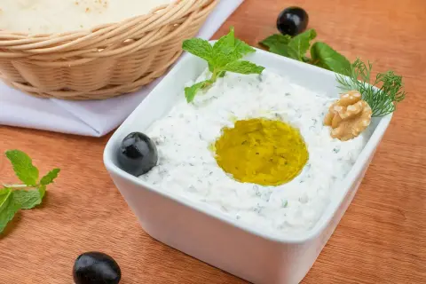 Jak zrobić tzatziki? Kremowy dip jogurtowy z oliwą, miętą, orzechami i oliwkami. Obok koszyk z pitą.