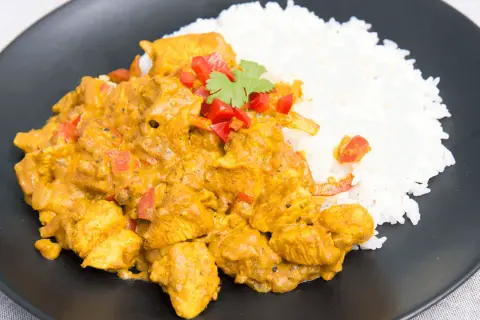 Aromatyczny kurczak curry z ryżem, udekorowany papryką i kolendrą, podany na czarnym talerzu.