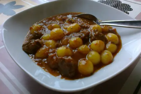 Bogracz z kluseczkami rączka gotuje. Aromatyczny gulasz z mięsem i ziemniakami w sosie, podany w białej misce.