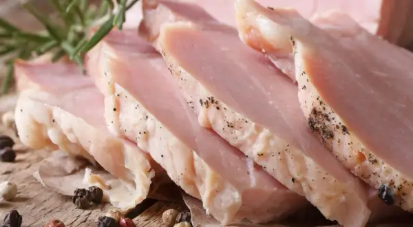 Super delikatny i soczysty schab gotowany, pokrojony w plastry, z gałązką rozmarynu i ziarnami pieprzu na drewnianym tle.