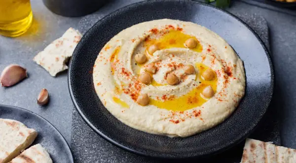 Kremowy hummus na talerzu, polany oliwą, posypany papryką i ziarnami ciecierzycy. Obok kawałki pity i czosnek.