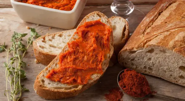 Nduja co to? Pikantna, pomarańczowa pasta na chlebie, obok świeżego pieczywa, papryki i ziół.