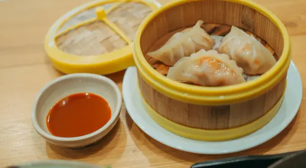 Parujące pierogi gyoza w bambusowym parowniku, obok miseczka z sosem.