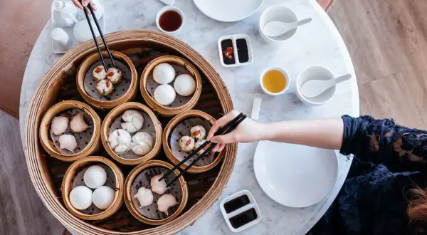 Ręce sięgają po pyszne dim sum z bambusowego parownika. Na stole stoją miseczki z sosami i herbatą.