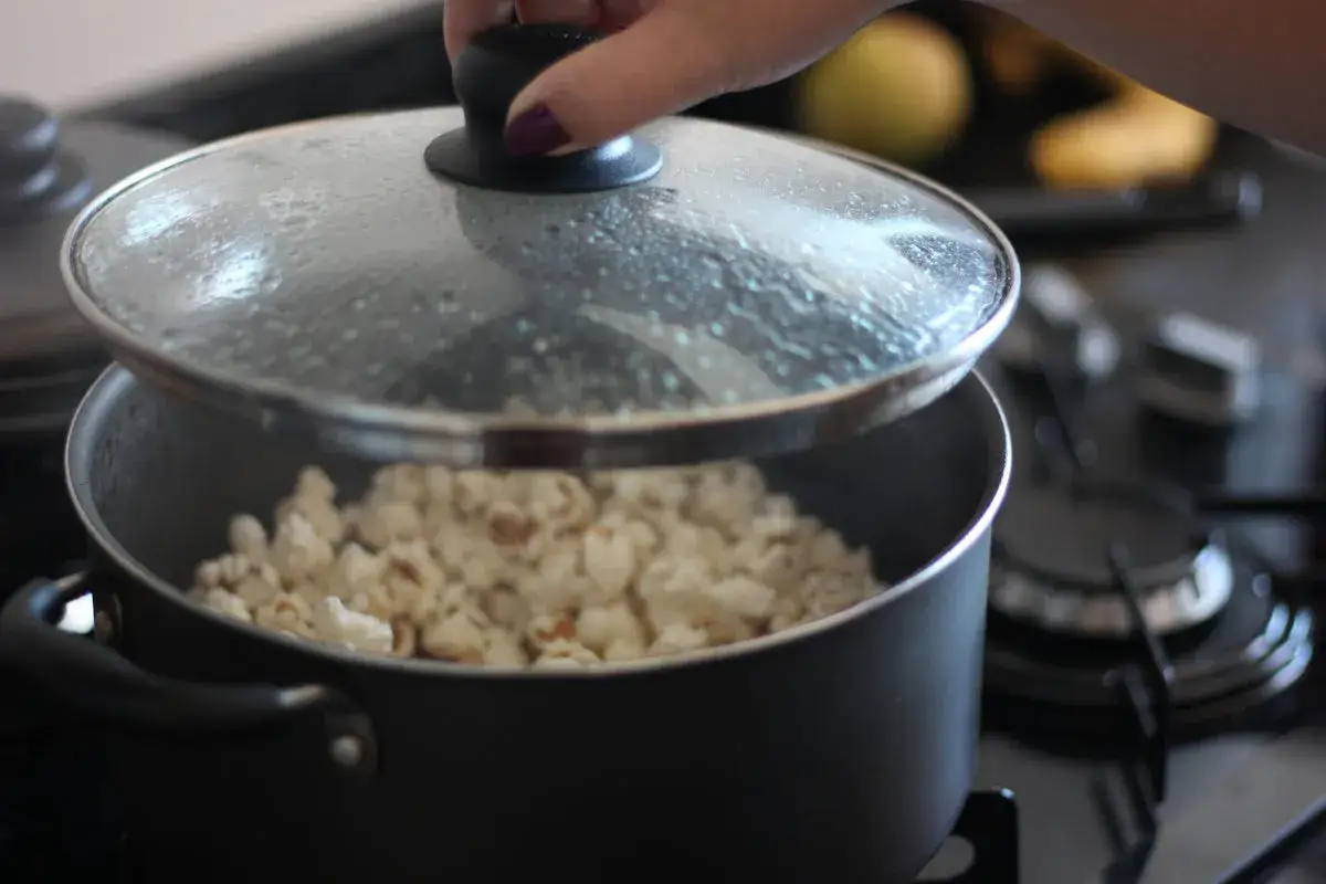 Ręka podnosi pokrywkę garnka, spod której widać gotowy popcorn. Prosty sposób, jak zrobić popcorn w garnku.