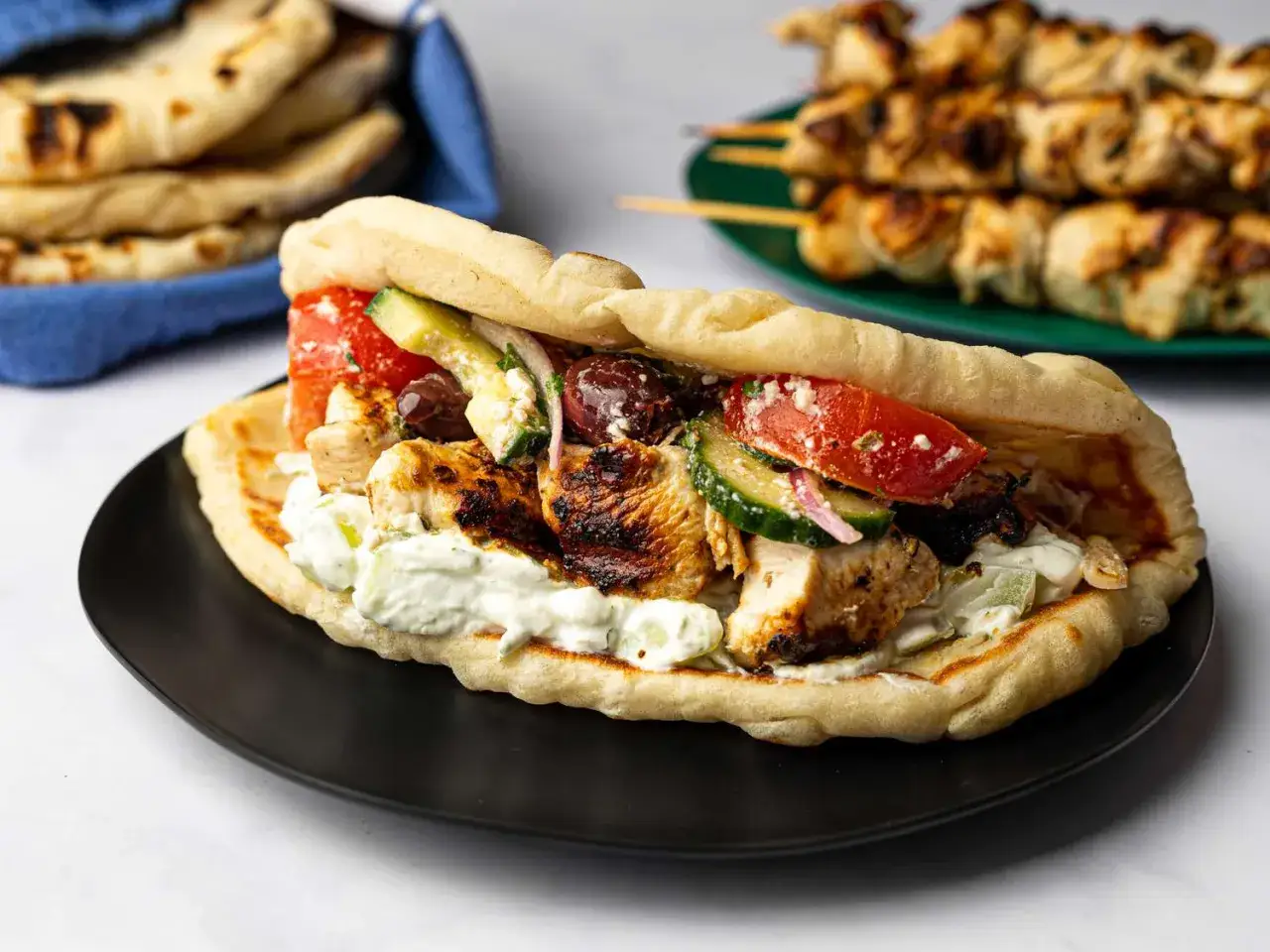 Pyszne souvlaki z kurczaka w picie z pomidorami, ogórkiem, fetą i sossem tzatziki. W tle widoczne szaszłyki.