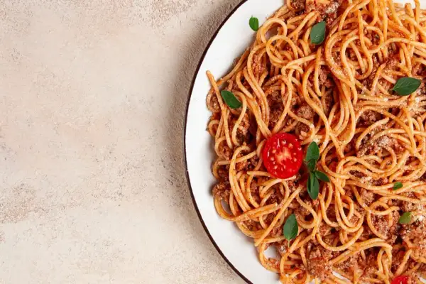 Talerz spaghetti bolognese z pomidorkiem i bazylią na jasnym tle.