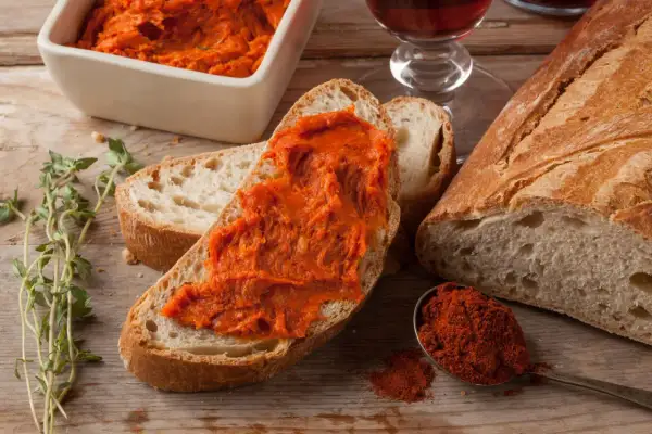 Nduja co to? Pikantna, pomarańczowa pasta na chlebie, obok świeżego pieczywa, papryki i ziół.