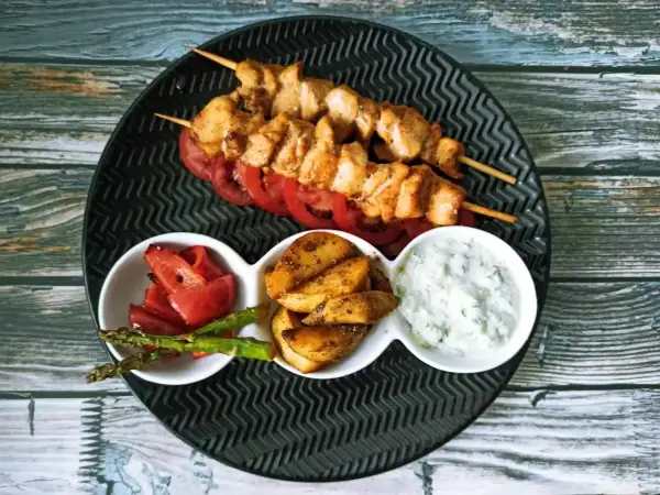 Dwa szaszłyki z kurczaka, czyli pyszne souvlaki, podane z pieczonymi ziemniakami, papryką, szparagami i sosem tzatziki.