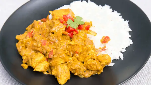 Aromatyczny kurczak curry z ryżem, udekorowany papryką i kolendrą, podany na czarnym talerzu.