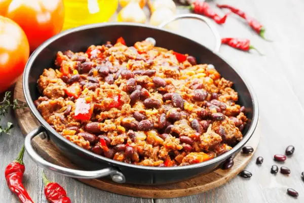 Pyszne chili con carne przepis w głębokiej patelni, z fasolą, mięsem mielonym i papryką. Obok pomidory i papryczki chili.